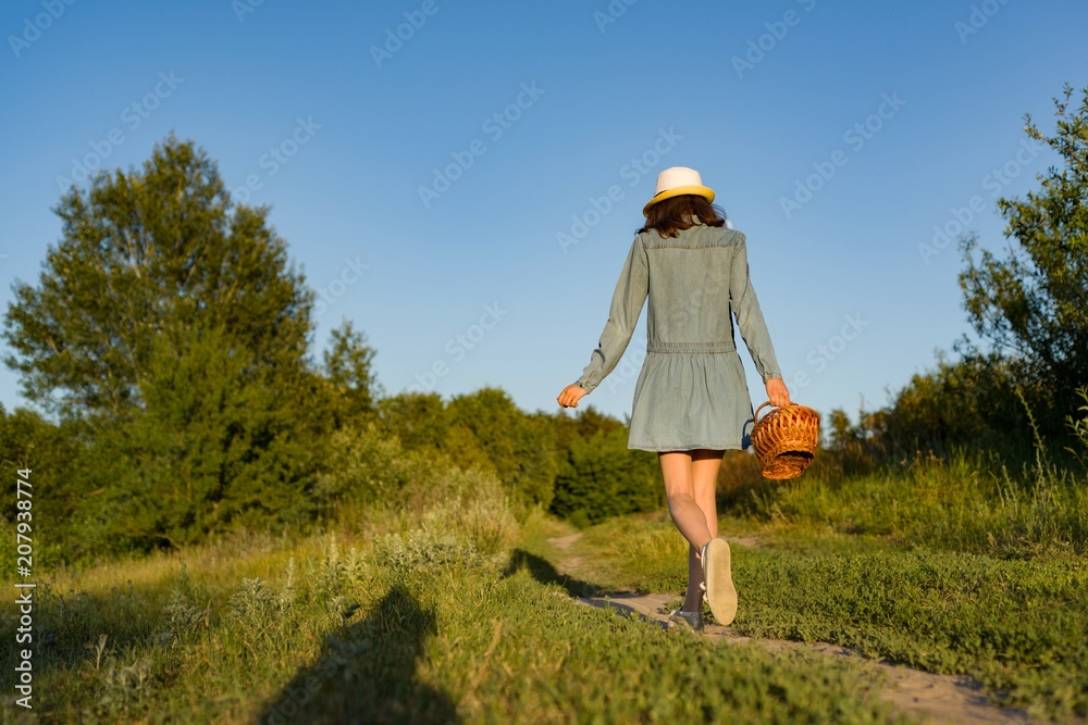 Outdoor summer portrait of teen girl with basket strawberries, straw hat. A girl on country road, back view.