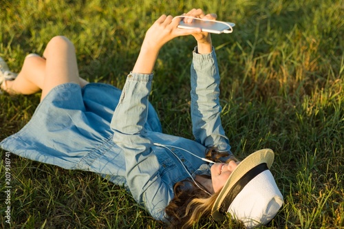 Portrait of teenage girl 14 years old lying on the grass. Girl in dress hat, in her headphones holds a smartphone, listens to music makes a selfi photo photo