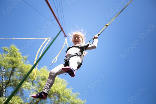 Child get fun on children trampoline with safety ropes. Little girl jumping high on blue sky background. Children summer entertainment.