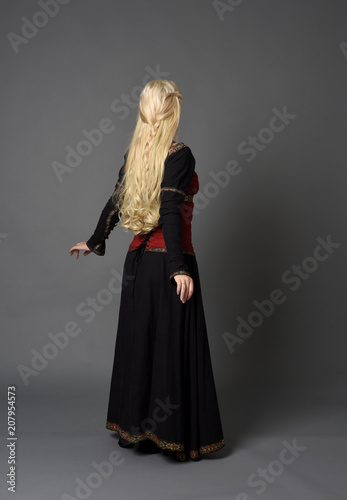 full length portrait of pretty blonde lady wearing  a red and black fantasy medieval gown. standing pose on grey background.