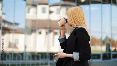 Young lady walks with a cup of coffee along the mirror wall outside and types something in her smartphone photo