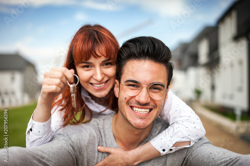 Smiling couple with keys to their new home hugging and looking at camera taking selfie