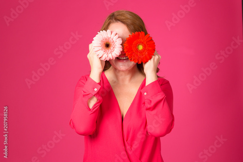 Joy red-hair woman, covering her eyes with a flower. Beautiful adult woman with a gerbera in hands on a pink background.