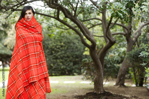 Giovane donna si riscalda con una coperta rossa avvolta intorno al corpo - sfondo vegetazione e alberi di un parco