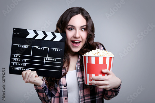 Young beautiful woman holding bucket with popcorn and clapper board on grey background