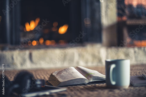 Shallow depth of field photo of open Bible, coffee, notepad and tablet in front of fire place