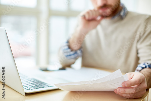 Faceless shot of man sitting with laptop at table and reading through papers in daylight. 