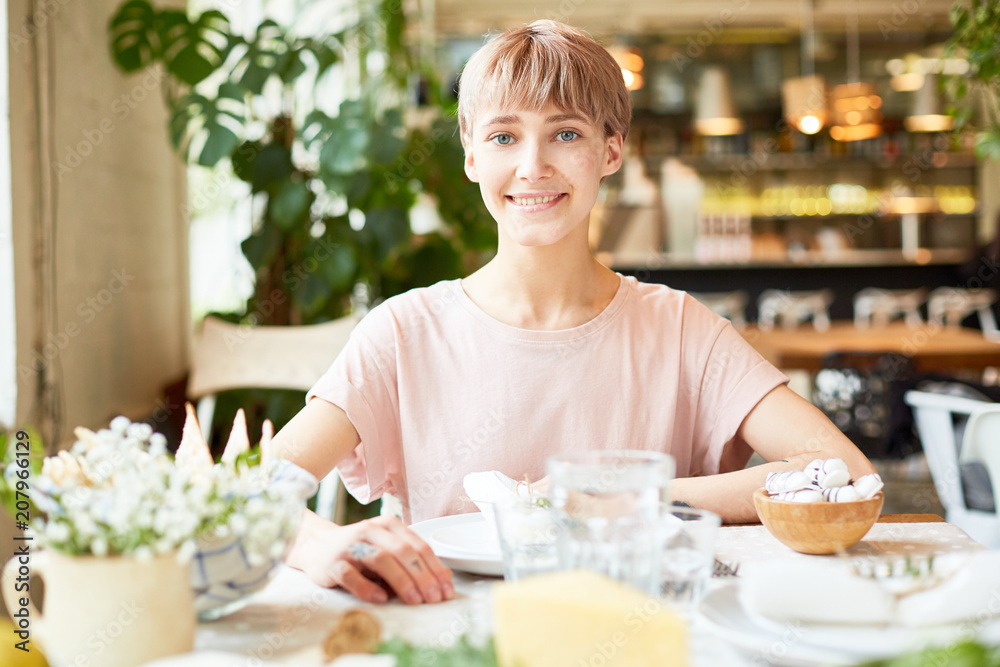 Beautiful young woman with short haircut sitting at table smiling and looking at camera on background of blurred restaurant