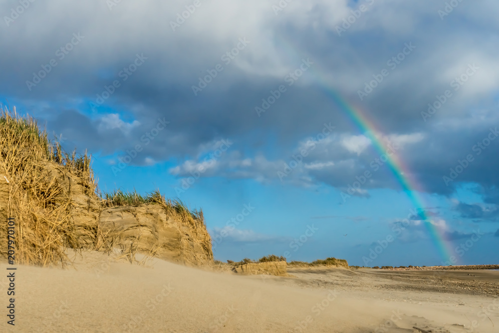 Sand dunes at a beach with a blue sky and grey clouds lead to a colorful rainbow in the back.