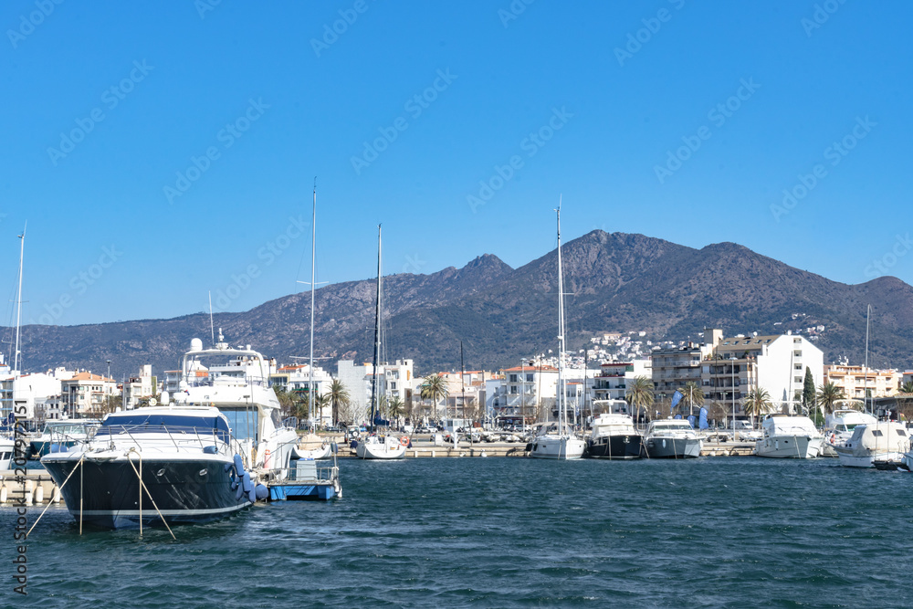 Motor and sailboats ancoring in the marina in Roses, Spain. The water is colored deep blue, while the sky has a lighter tint. In the back the start of the pyrenees can be seen