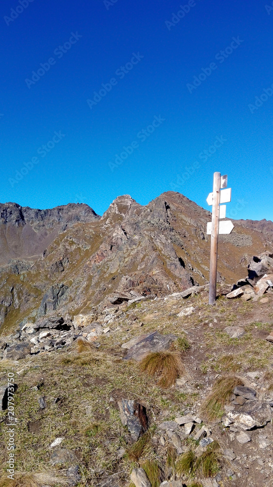 Crossroad signpost on a mountain peak at the end of an hiking path (Italian Alps landscape). Photo taken in a sunny summer day. Clear blue sky in the background. 