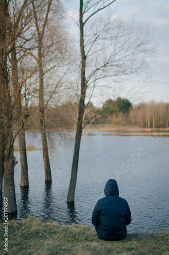A guy in a jacket with a hood on his head sits on the river bank