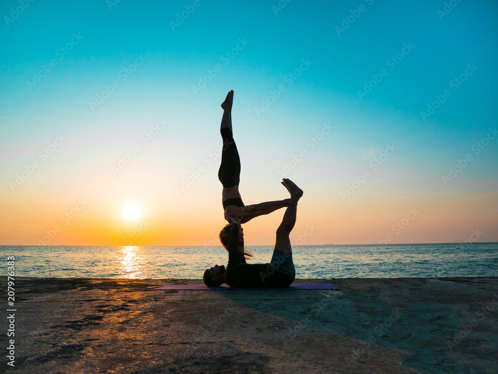 Silhouettes of fit young couple doing acro-yoga at sea beach. Man lying on concrete  plates and balancing woman in his feet. Beautiful couple practicing yoga  together Stock Photo