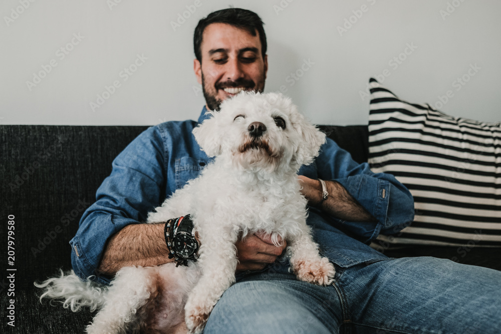 Handsome young man playing with his cute little white dog at home. Relaxed on the sofa. Lifestyle. Pet photography.