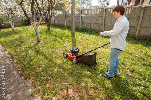 Young man in sunglasses, jeans and shirt mows the grass on the lawn in the village in the summer.