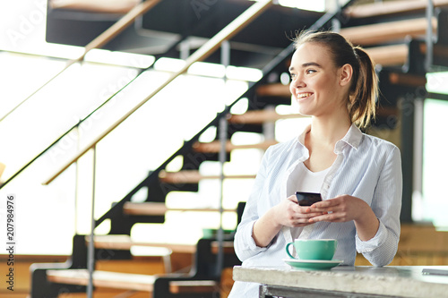 Pretty young girl standing with cup of coffee on counter and holding smartphone while laughing away photo