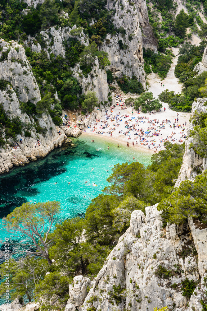 View from above of the calanque of En-Vau, a hard-to-reach narrow natural creek with white sandy beach close to Marseille and Cassis, with people sunbathing and swimming in the crystal clear water. View from above of the calanque of En-Vau, a hard-to-reach narrow natural creek with white sandy beach close to Marseille and Cassis, with people sunbathing and swimming in the crystal clear water.