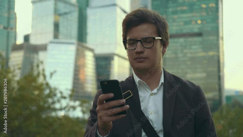 Handsome young businessman in glasses and a suit swiping across his smartphone screen walking in Moscow city. Daylight. Skyscrapers. Tracking medium shot Handsome young businessman in glasses and a suit swiping across his smartphone screen walking in Moscow city. Daylight. Skyscrapers. Tracking medium shot