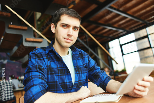 Handsome young man in casual blue shirt holding tablet and sitting at desk with studies looking at camera