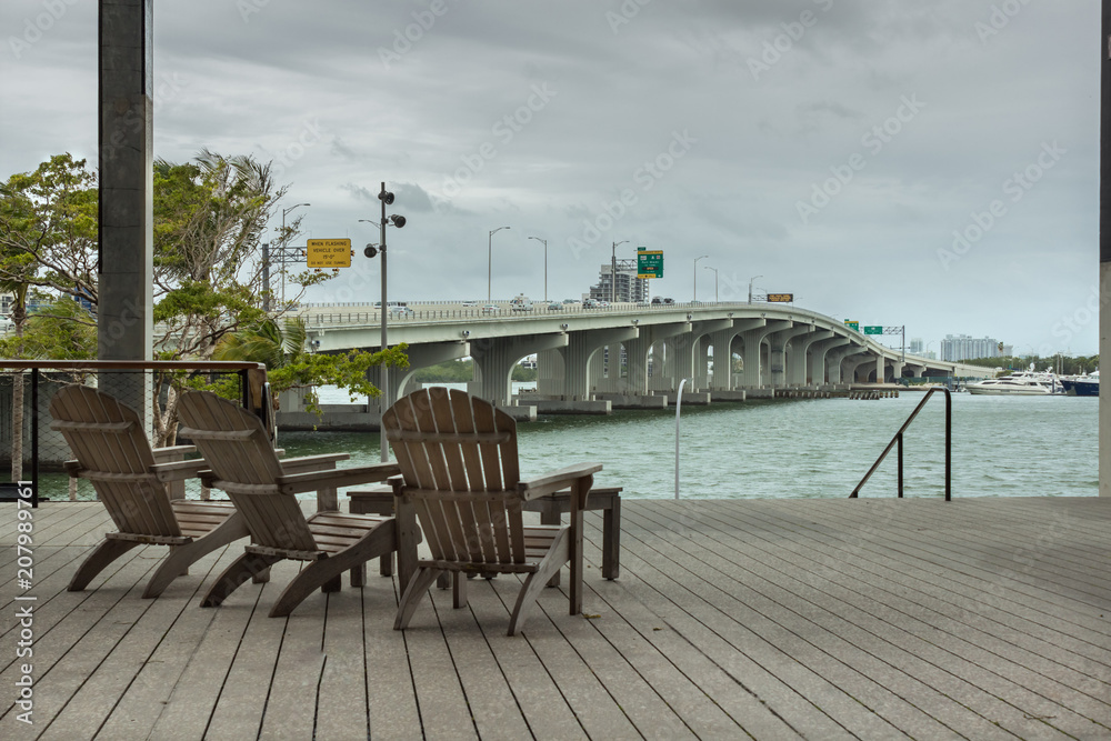 Sillas en una terraza de madera con vista al mar y al puente con cielo nublado