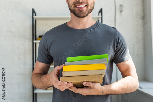 cropped image of smiling man standing with stack of books at home