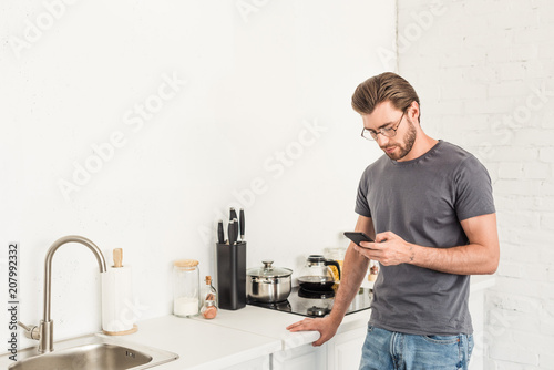 front view of young man in eyeglasses checking smartphone at kitchen