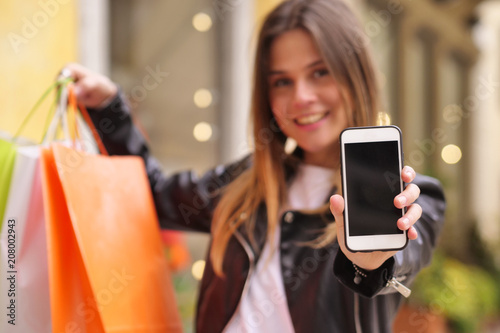Beautiful young woman (girl) shows her smartphone and shopping bags with the city in the background.
