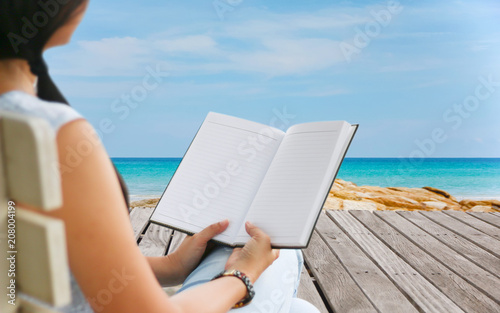 Rear view of woman reading novel by the sea.Summer beach background. Sand and sea and sky