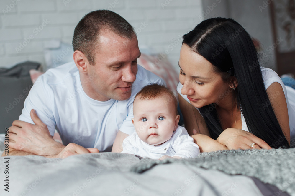 Young mom and dad and their baby son lie in bed and look at the camera indoors. The concept of a happy family