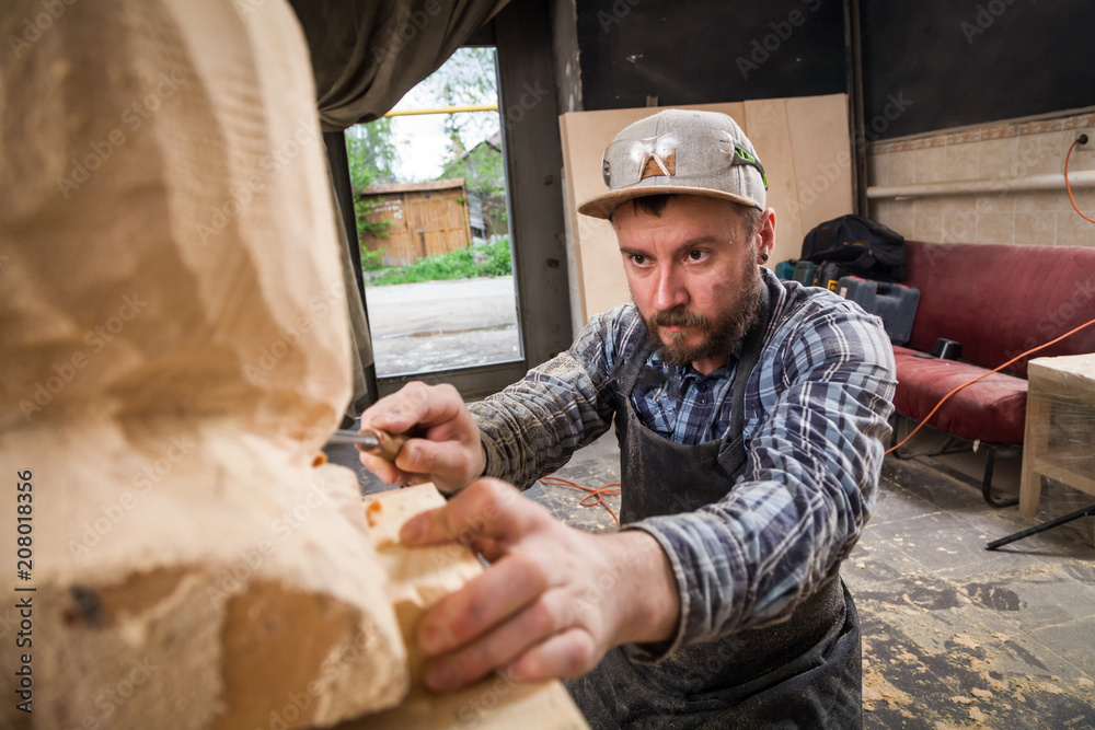 Young man carpenter in a work clothes saws a man's head with a tree , using a chisel in the workshop, around a lot of tools for work Young man carpenter in a work clothes saws a man's head with a tree , using a chisel in the workshop, around a lot of tools for work