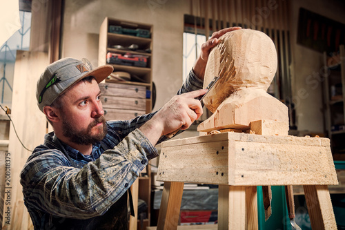 Young man carpenter in a work clothes saws a man's head with a tree , using a chisel in the workshop, around a lot of tools for work