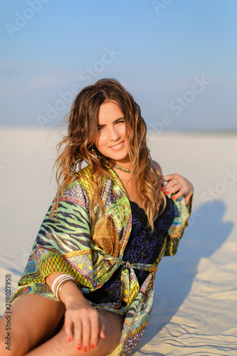 Young beautiful woman wearing green beach robe and having red nails sitting on sand. Concept of beach photo session and summer.