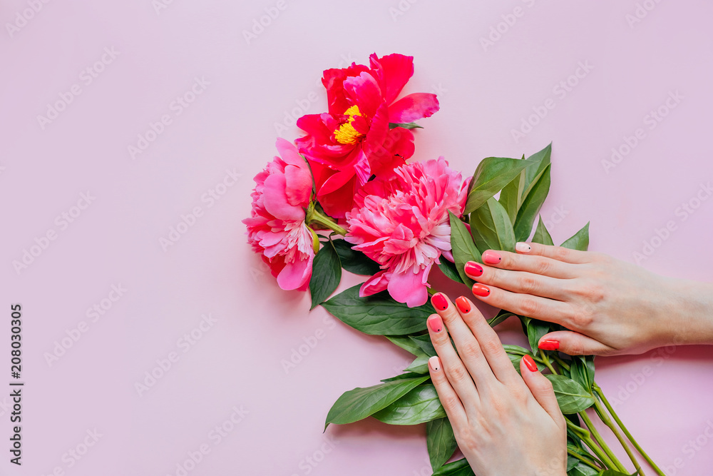 Creative bright trendy summer manicure with nails of different color. Two female hands with art nail design on pink background and fuchsia peony flower bouquet.