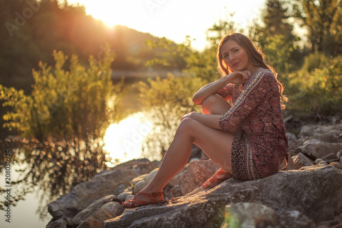 Young woman, wearing dress, sitting on stones by the river with golden sunset light in background.