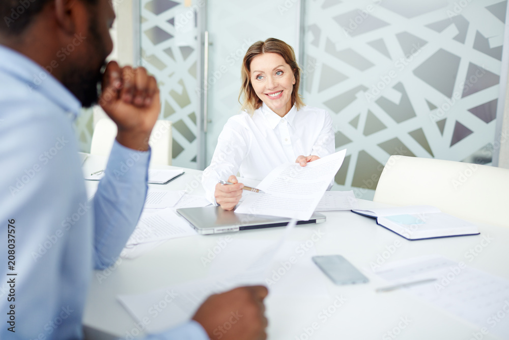 Female employee in elegant clothes holding contract at table with black confrere in light office Female employee in elegant clothes holding contract at table with black confrere in light office