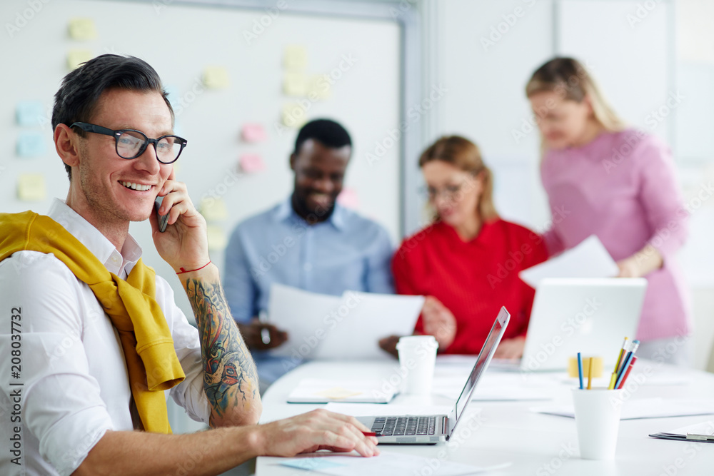 Man in shirt with tattoo and glasses at table talking on phone and using laptop with papers