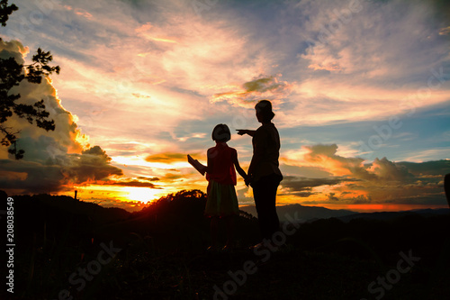 Silhouettes of mother and douther looking sunset on peak of mountain.