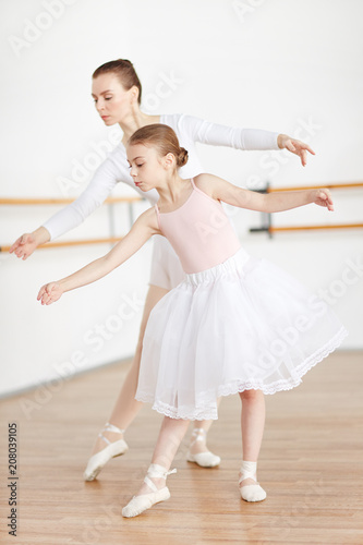 Woman and girl with strict hairstyle in light pointe and tutu staying in poses in white studio 