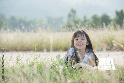 A little girl is lying on her stomach reading on the grass. She has a look of enjoyment on her face and she looks very relaxed. There is quite a bit of negative space around her.