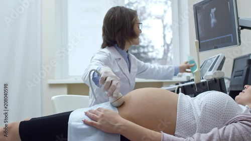 Happy pregnant woman lying on examination coach, talking and looking at monitor while smiling female doctor applying gel to ultrasonic transducer and doing test
