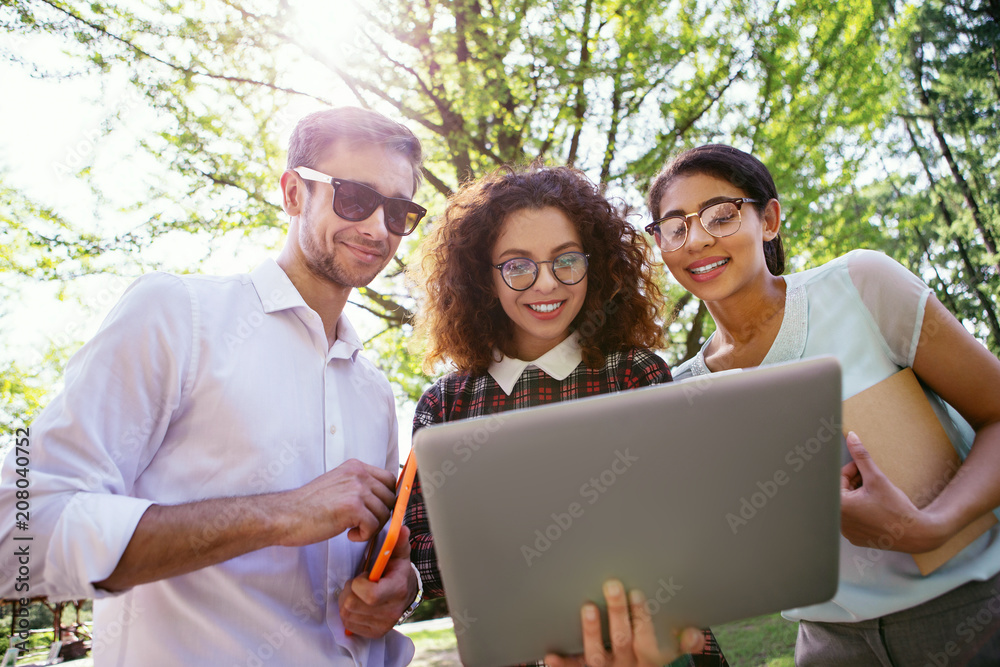 Pleasant studying. Joyful curly-haired lady holding her laptop and her friends standing near her