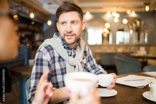 Handsome man in casual outfit drinking hot beverage and listening to colleague while sitting at cafe table together.