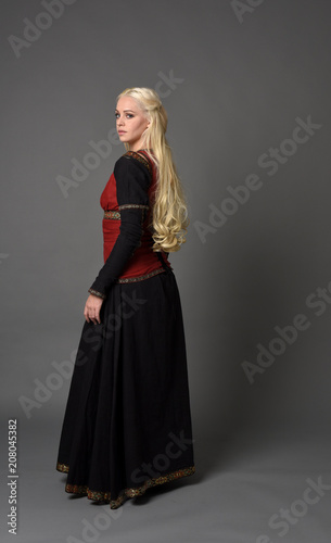 full length portrait of pretty blonde lady wearing  a red and black fantasy medieval gown. standing pose on grey background.