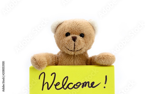 Brown teddy bear holding with the two hands a note in bright yellow color with the handwritten message “Welcome!” as welcome sign concept. Photo isolated in a seamless white background. © fewerton