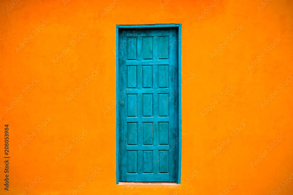 Vivid bright orange colour facade with blue-green closed door in the center of large empty orange wall texture background space. Vivid bright orange colour facade with blue-green closed door in the center of large empty orange wall texture background space.