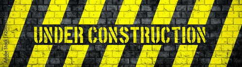 Under construction warning sign with yellow and black stripes on concrete wall texture background in wide panorama format. Concept for do not enter the area, caution, danger, construction site.