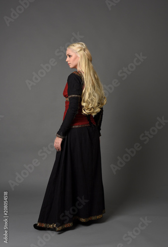 full length portrait of pretty blonde lady wearing  a red and black fantasy medieval gown. standing pose on grey background.