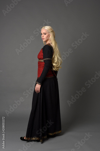 full length portrait of pretty blonde lady wearing  a red and black fantasy medieval gown. standing pose on grey background.