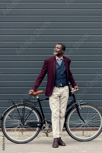 stylish mature african american man in burgundy jacket posing near bicycle