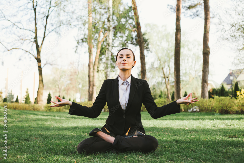 Beautiful concentrated business woman in a black suit sits in a lotus pose and meditates. Yoga outdoors Beautiful concentrated business woman in a black suit sits in a lotus pose and meditates. Yoga outdoors